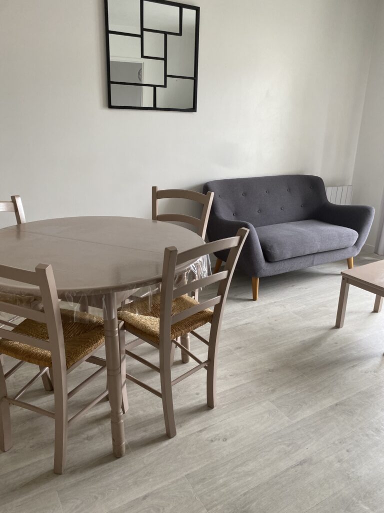 Pièce de vie avec table ronde en bois, chaises assorties et canapé gris confortable sur parquet clair.