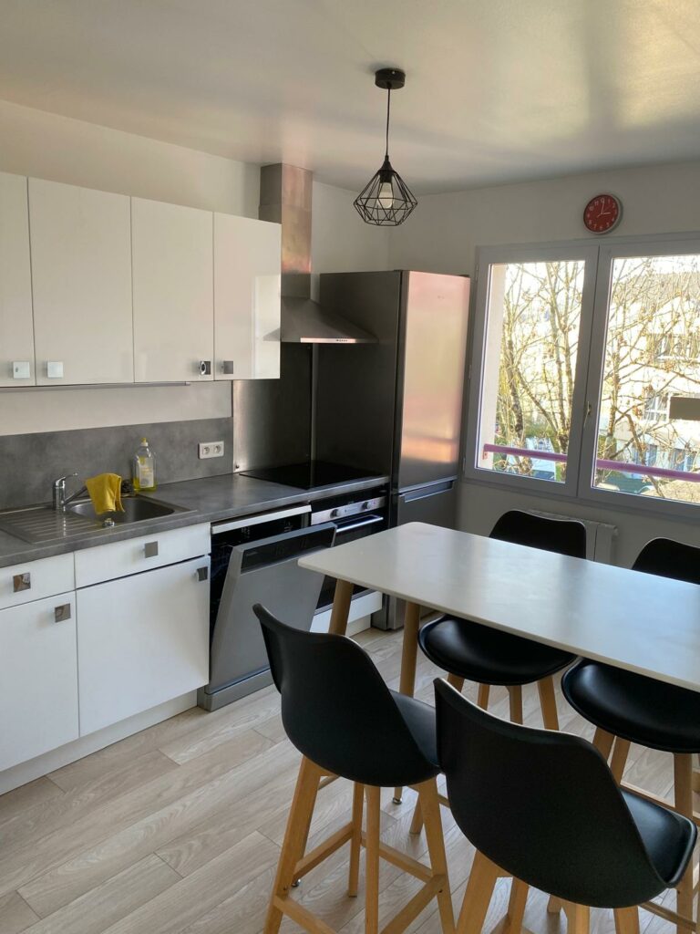 Cuisine équipée moderne avec façades blanches, plan de travail gris et table haute avec chaises noires.